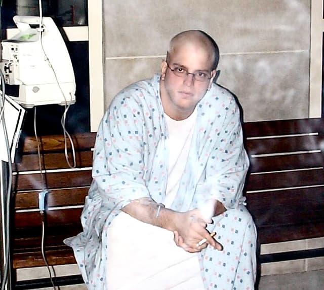 Rob Scott sitting on a hospital bench during cancer treatment, bald from chemotherapy, smoking under a No Smoking sign