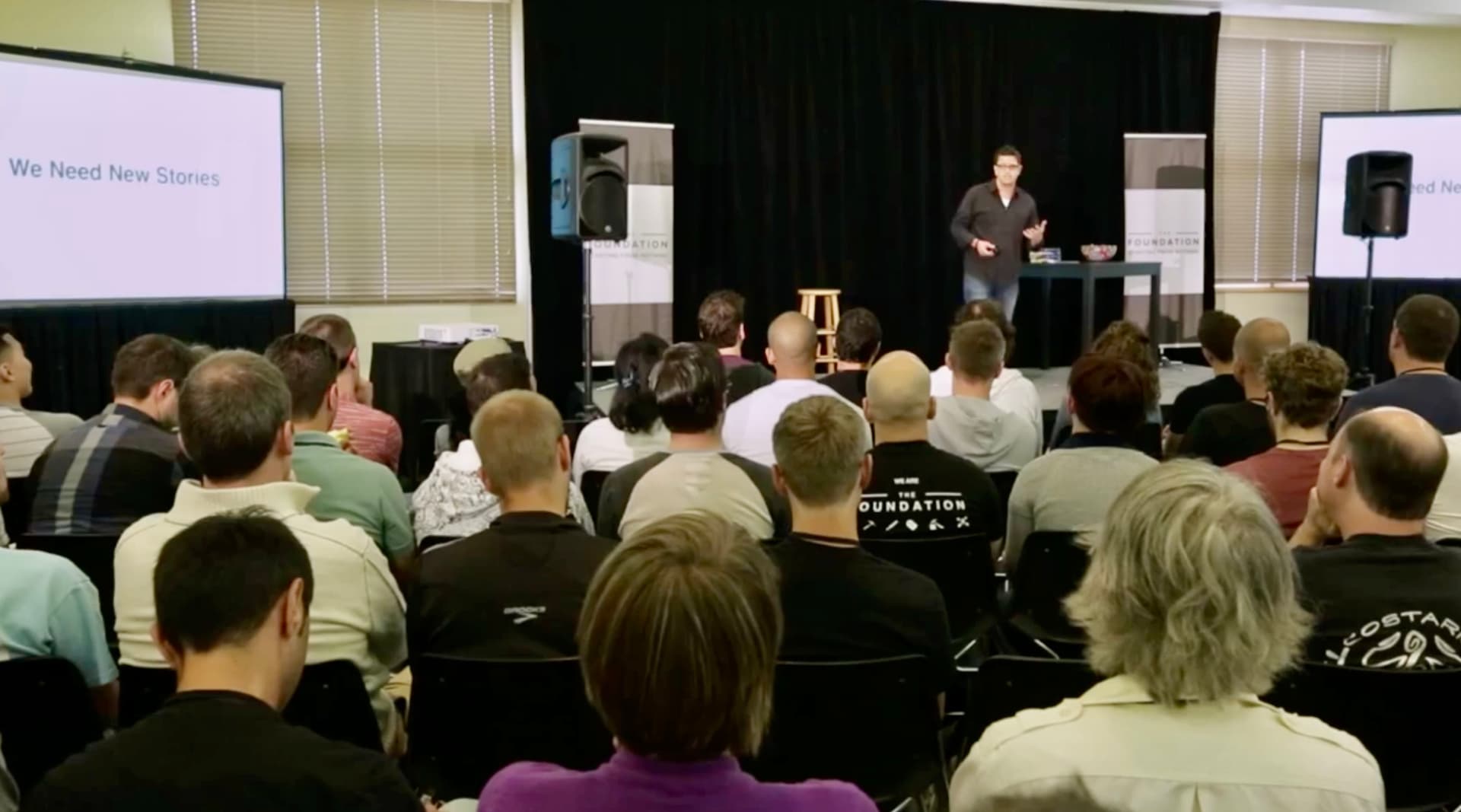 Rob Scott presenting to a large audience with 'We Need New Stories' on screen, demonstrating the scale and reach of Identity Shifting methodology